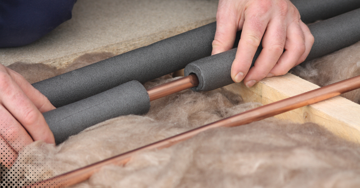 A person insulates copper pipes with foam tubing in an attic space.